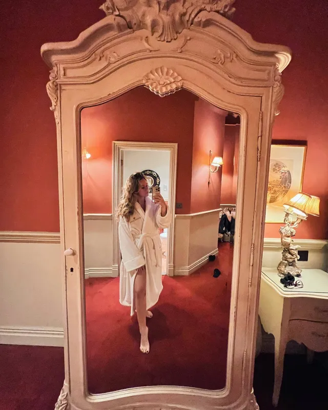 Brec Bassinger taking a mirror selfie in a pale dress and heels in an ornate room.