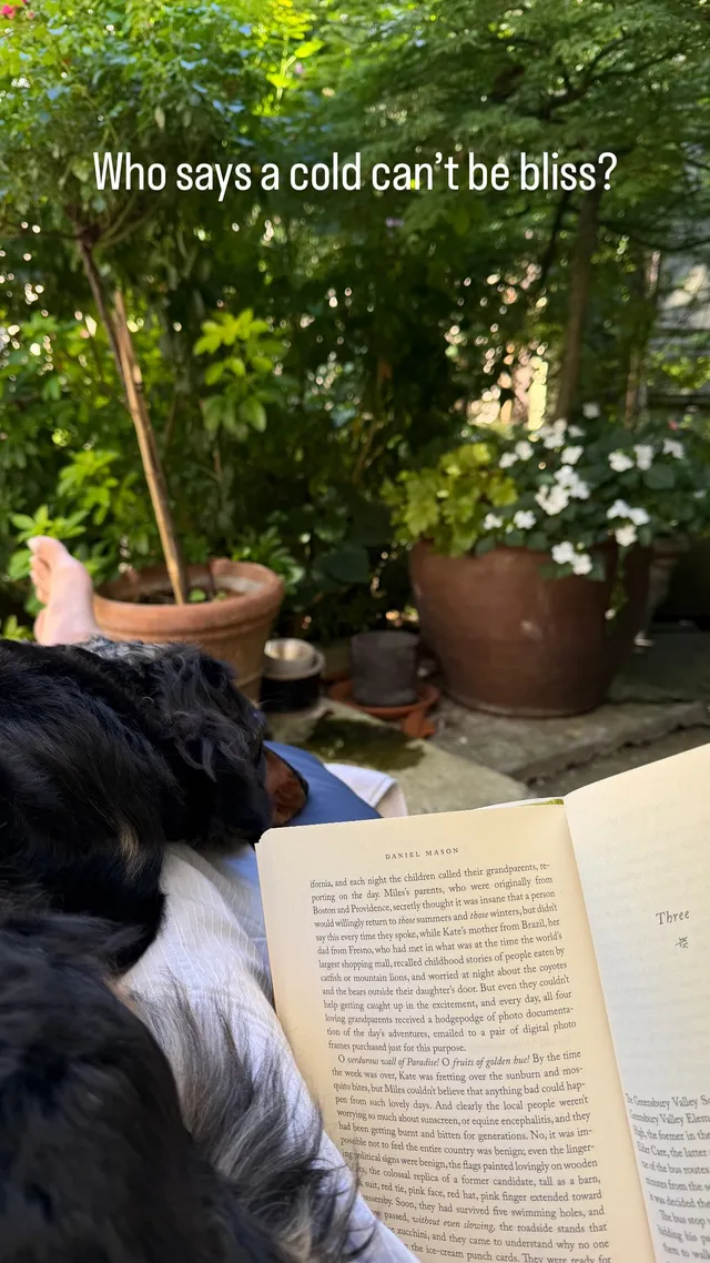 Emilia Clarke with bare feet extended beside an open book outdoors.