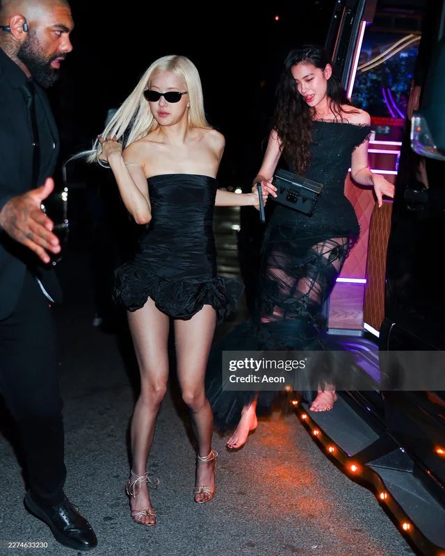 Rosé walking through a crowd in a strapless black dress and patterned heeled sandals.