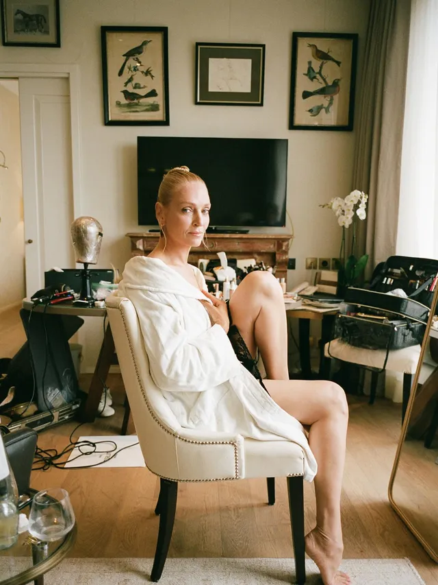 Uma Thurman sitting barefoot in a white outfit indoors.