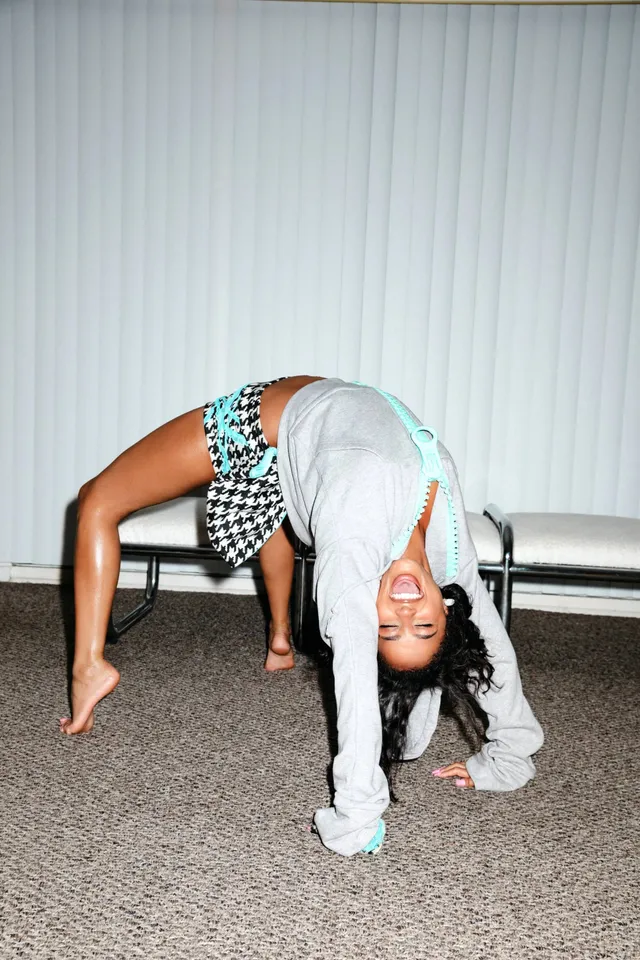 Tyla bending backwards barefoot beside a bench in a grey top and checked shorts.