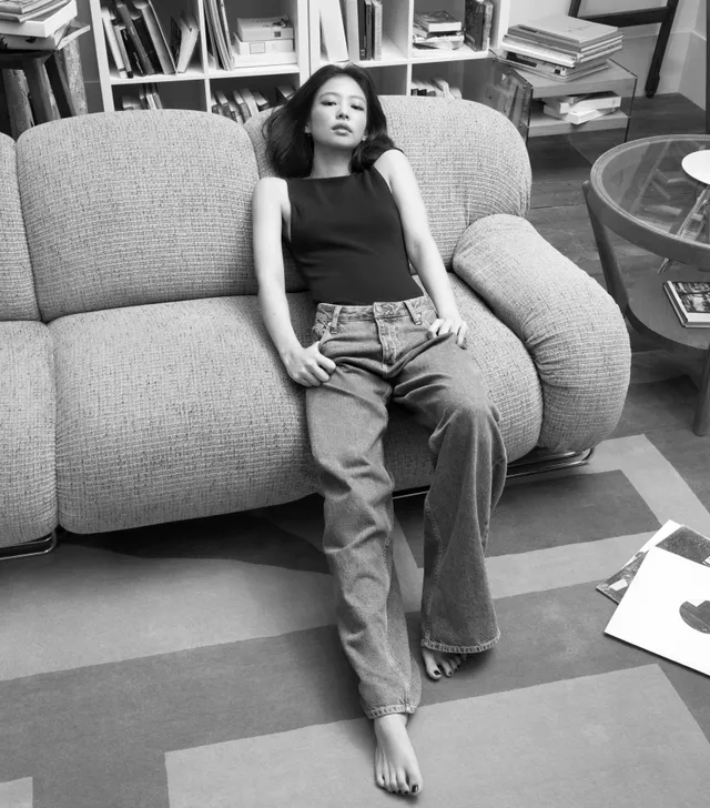 Jennie Kim sitting barefoot on a sofa in jeans and a dark top during a monochrome portrait.