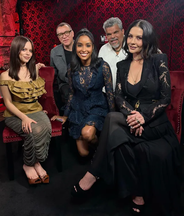 Catherine Zeta-Jones seated at a group event in a black dress and heels.