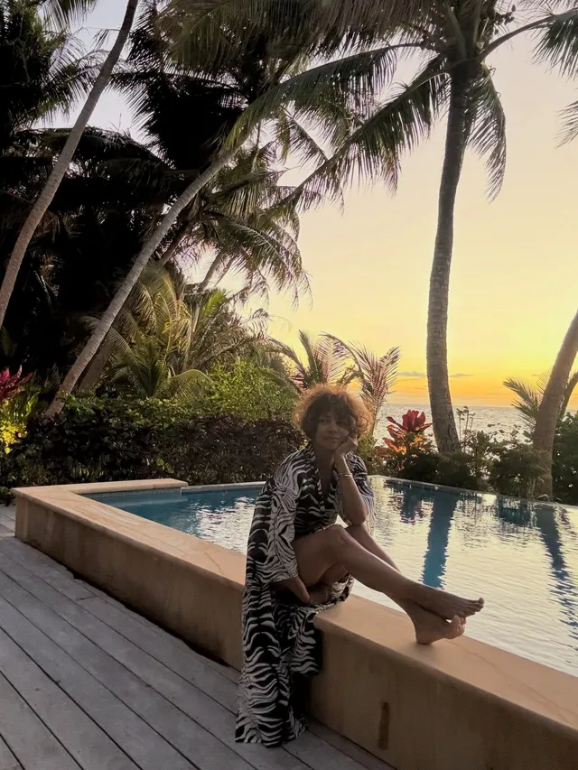 Halle Berry sitting barefoot beside a pool at sunset.
