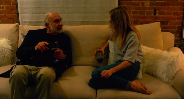 Sienna Miller sitting cross legged on a sofa in a screen scene.