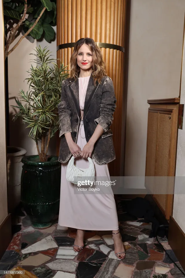 Dianna Agron posing at an event in a grey jacket and pale skirt.