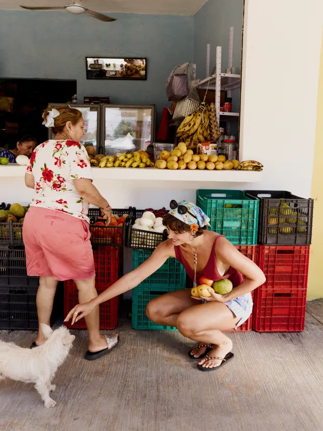 Taylor Hill crouching in flat sandals beside a dog at a produce stall.