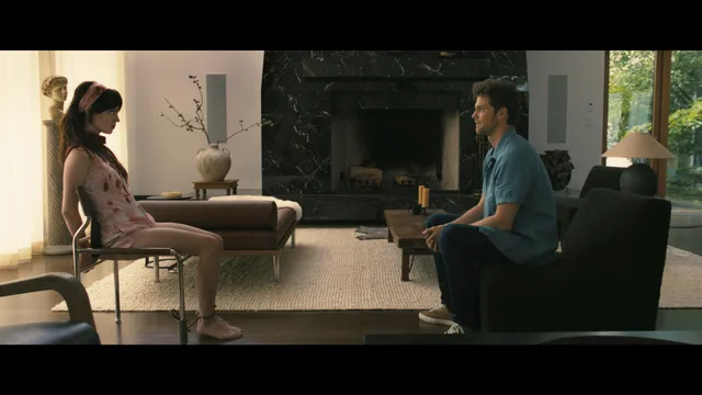 Sophie Thatcher sitting barefoot opposite a man in a bright indoor film scene.