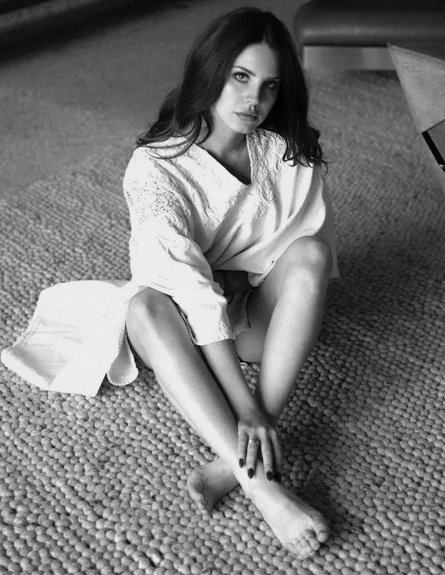 Lana Del Rey sitting barefoot on a carpet in a monochrome portrait.