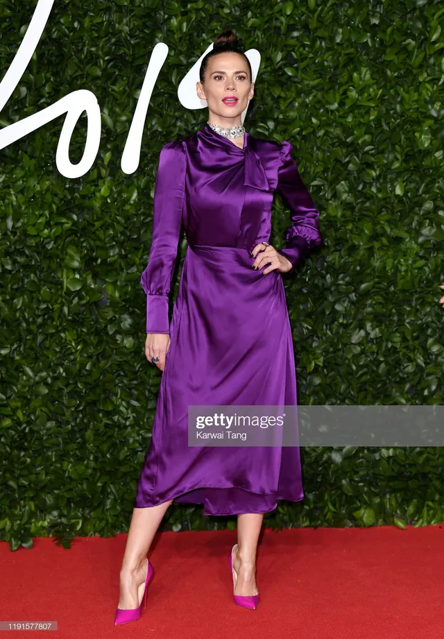 Hayley Atwell posing against a leafy wall in a purple dress.