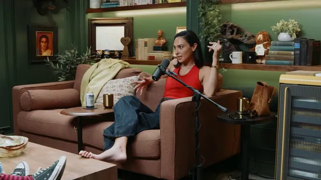 Gal Gadot sitting barefoot on a sofa during a conversation.