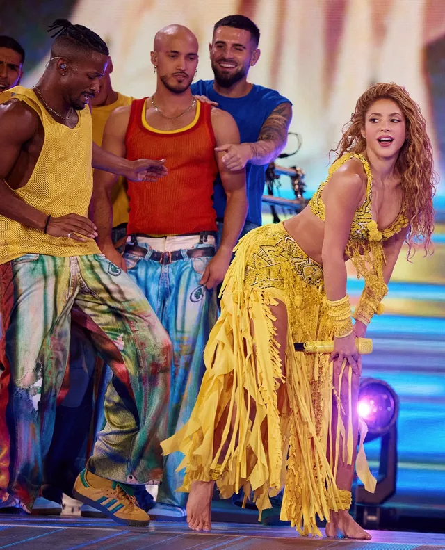 Shakira dancing barefoot beside performers in a yellow fringed outfit.