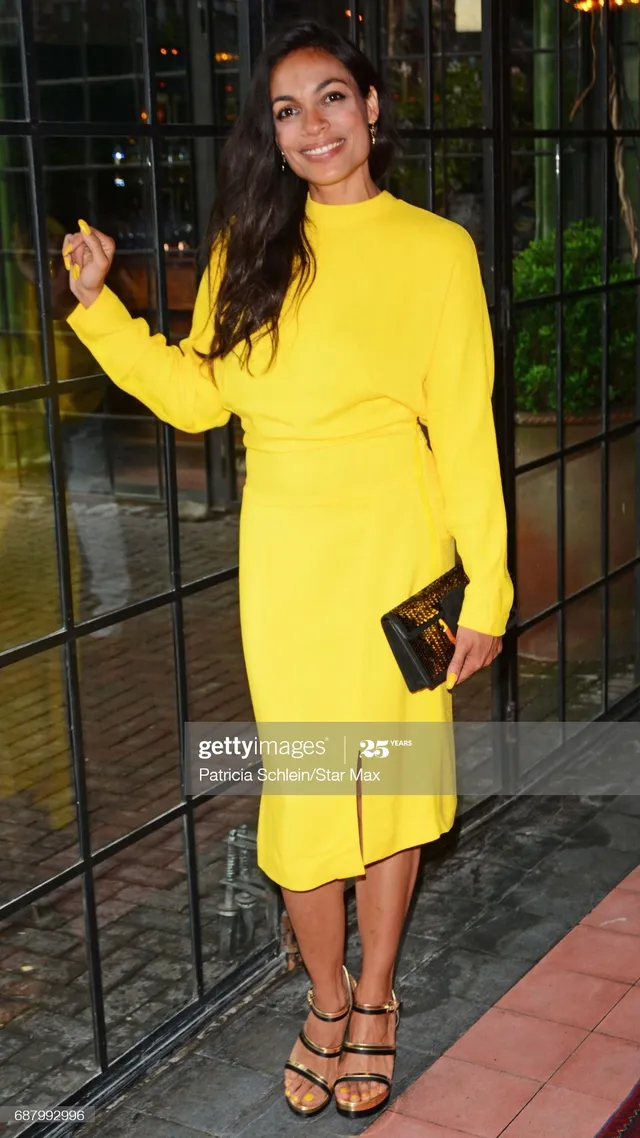 Rosario Dawson standing outside in a yellow dress and black sandals.
