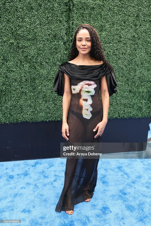 Tessa Thompson posing against greenery in a long black dress.