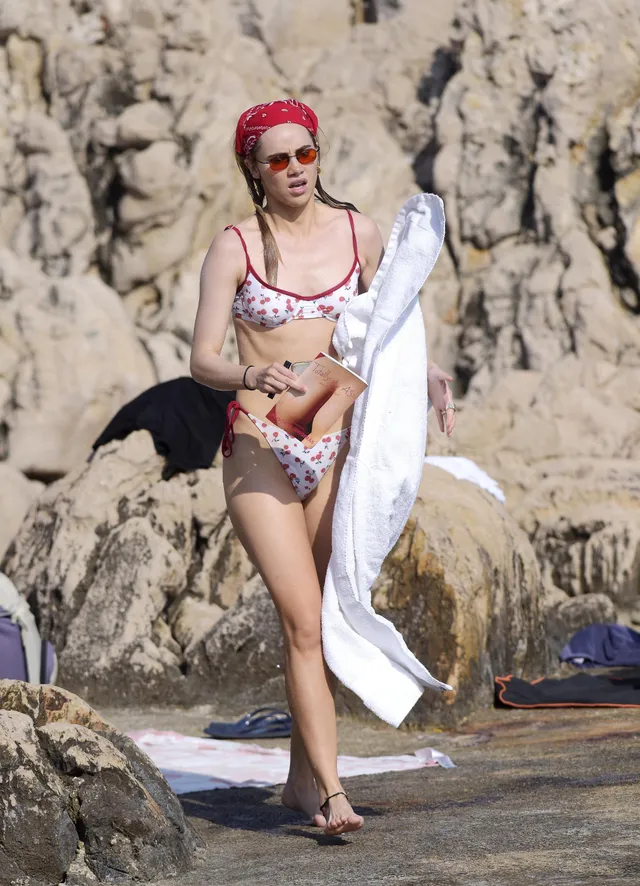 Suki Waterhouse standing by the sea holding a towel.