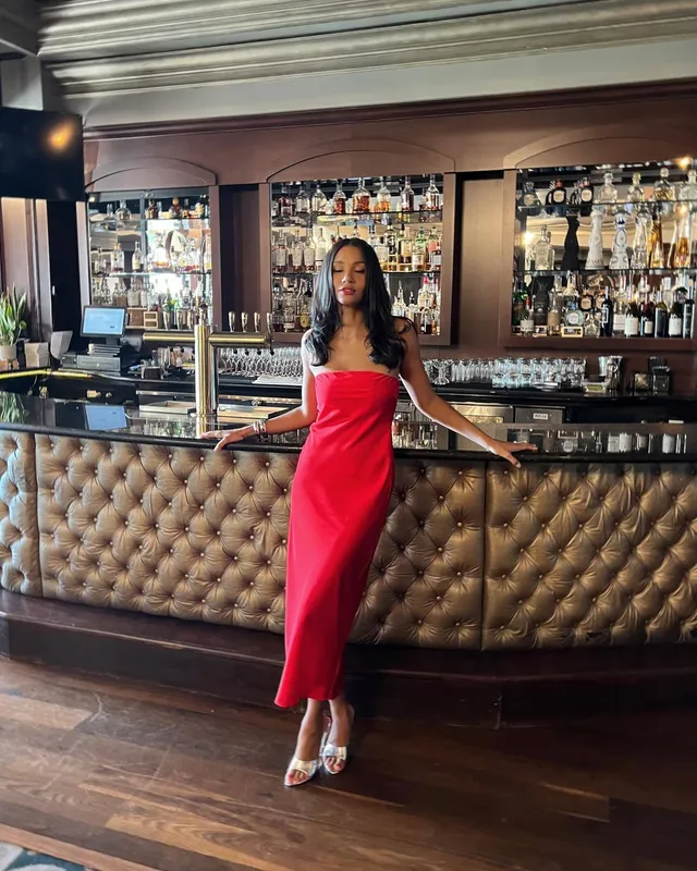 Greta Onieogou standing by a bar in a long red dress and clear heels.