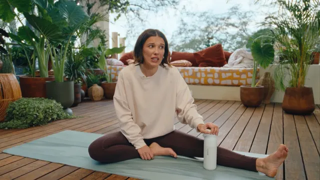 Millie Bobby Brown: seated barefoot on an exercise mat on wooden decking.