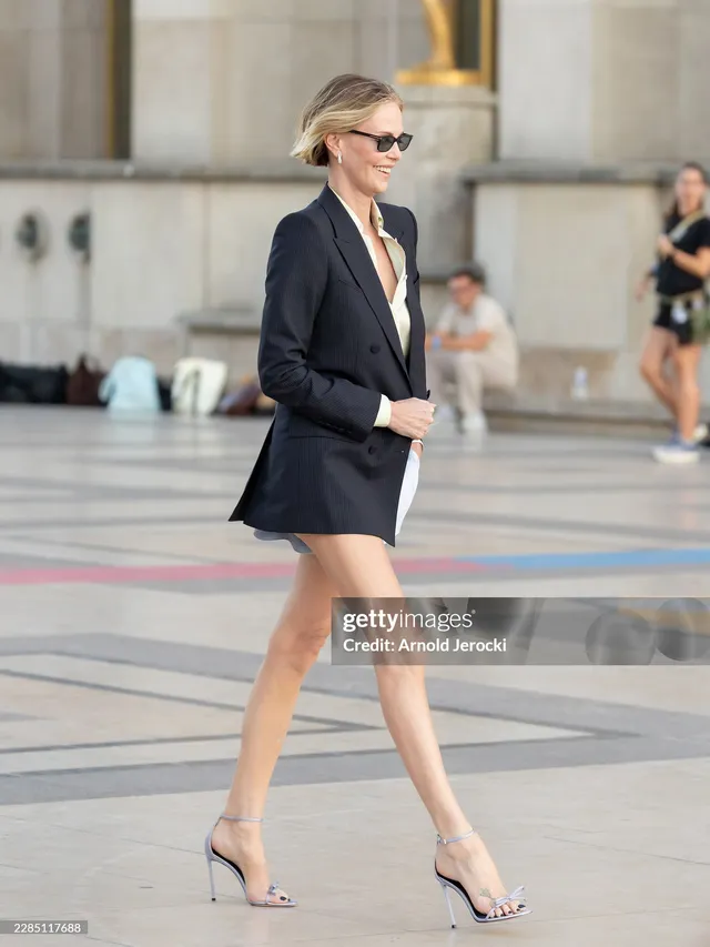 Charlize Theron walking outside in a black blazer and clear heels.