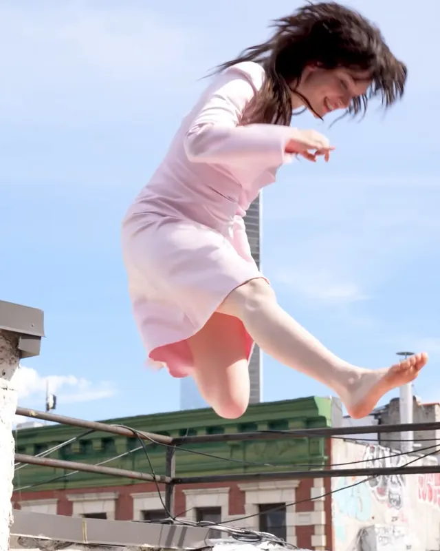 Sophie Thatcher jumping barefoot on a rooftop mattress in a pink dress.