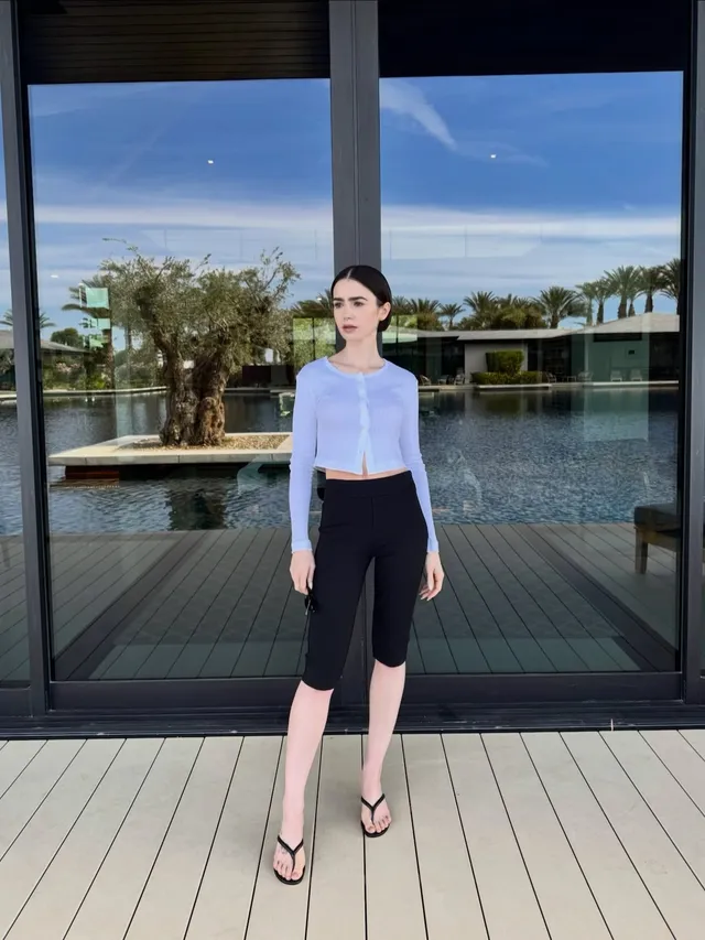 Lily Collins standing by water in cropped trousers and flip-flops.