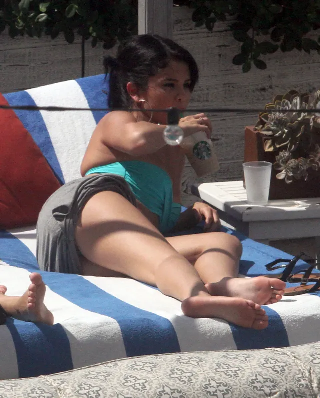 Selena Gomez reclining barefoot on a striped lounger beside a table.