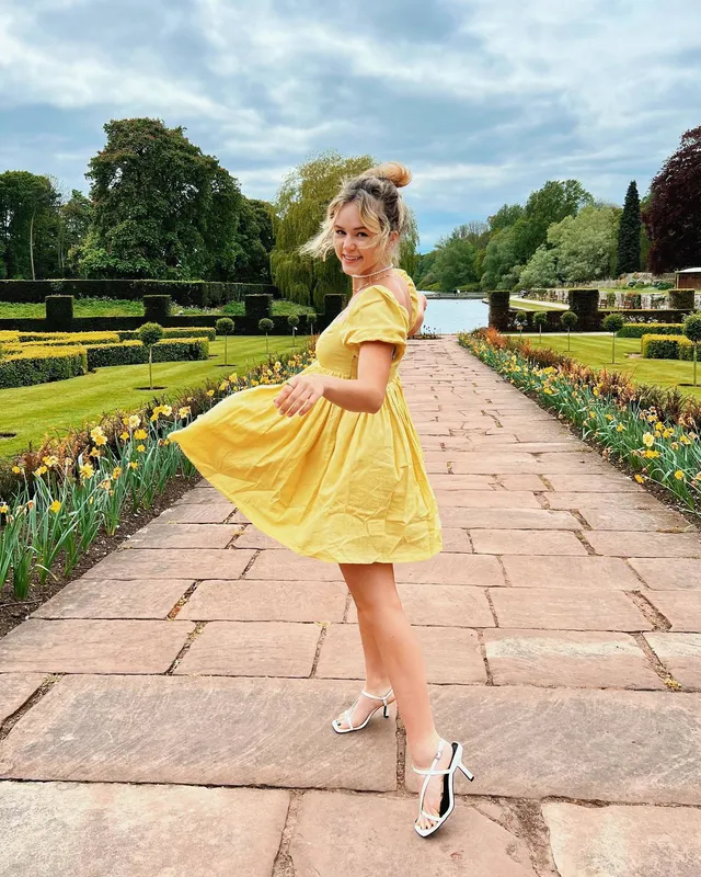 Brec Bassinger posing on a garden path in a yellow dress and white heels.