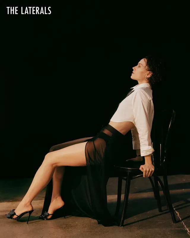 Daisy Ridley sitting on a chair in a white blouse and dark skirt.