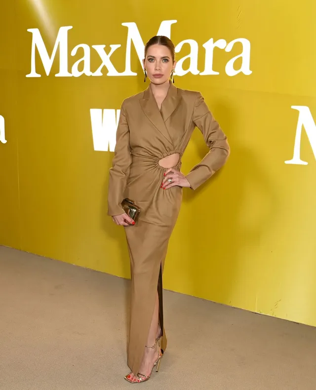 Ashley Benson standing at a fashion event in a tan dress and heels.