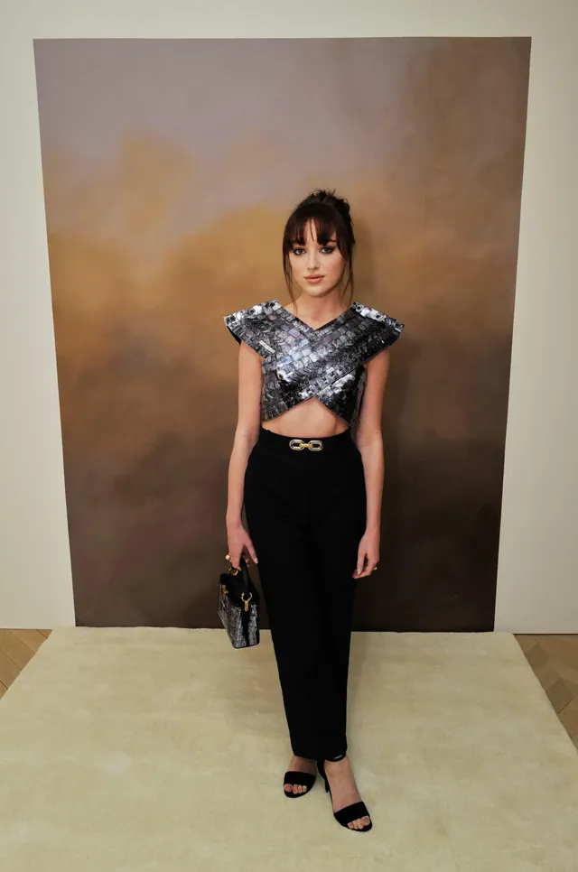 Phoebe Dynevor standing in a metallic top and dark skirt with heels.