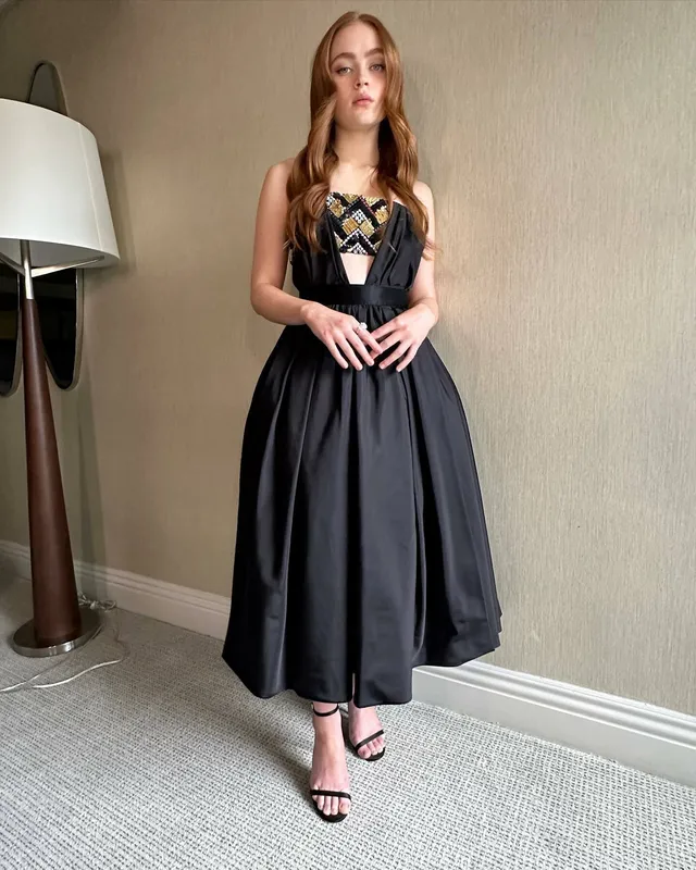 Sadie Sink standing in a black dress and heeled sandals indoors.