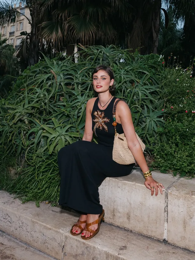 Taylor Hill sitting on a low wall wearing flat sandals and a dark outfit.