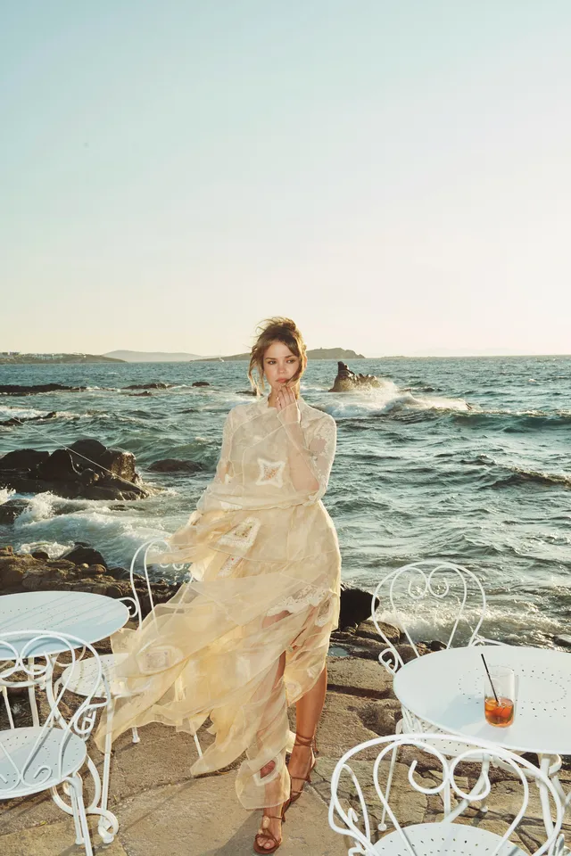 Maia Mitchell wearing a pale dress and heels by the sea.