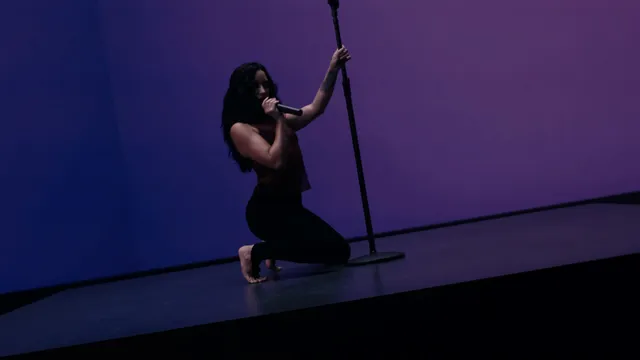 Demi Lovato kneeling barefoot beside a microphone in a purple-lit setting.