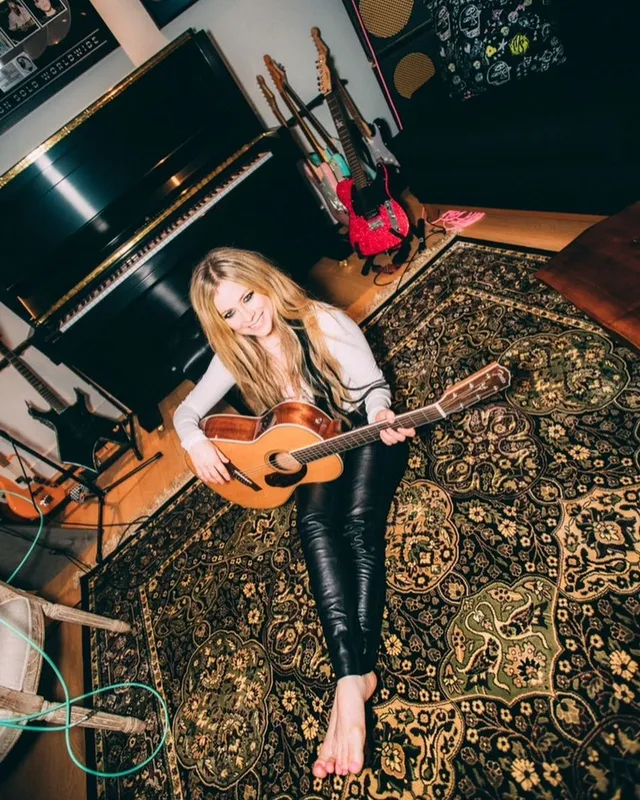Avril Lavigne sitting barefoot on a rug while playing guitar.