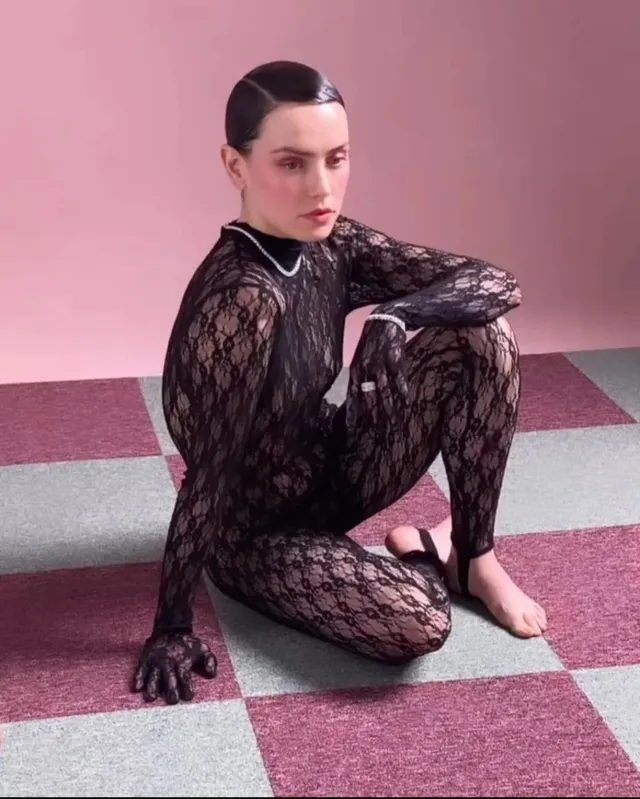 Daisy Ridley sitting on a patterned floor in a black lace outfit.
