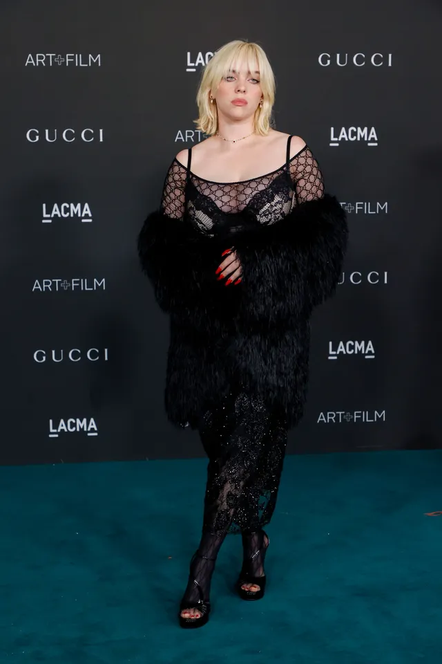 Billie Eilish posing at an event in a black lace dress and heels.