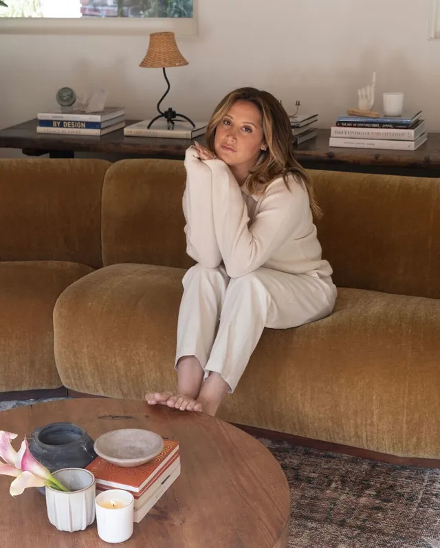 Ashley Tisdale sitting barefoot on a sofa in a pale knit outfit.
