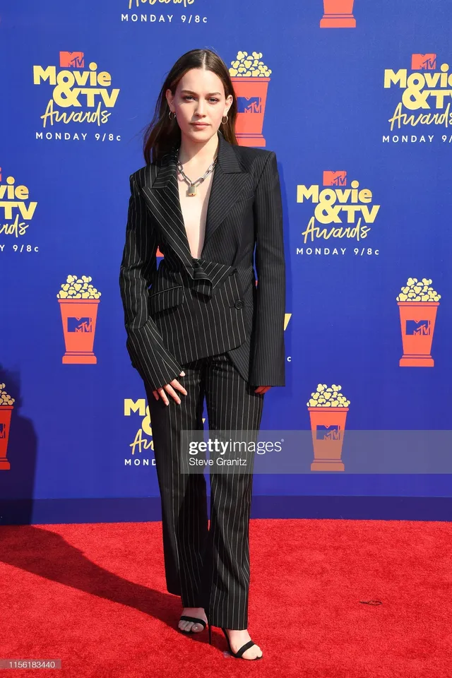 Victoria Pedretti standing on a red carpet in a dark suit.