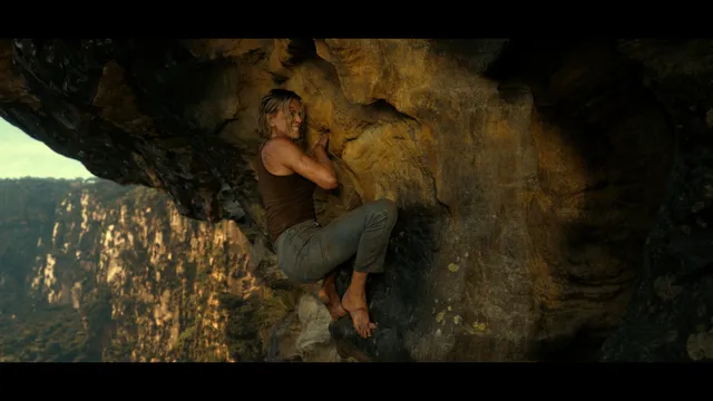 Charlize Theron a screen scene gripping a rock face barefoot.