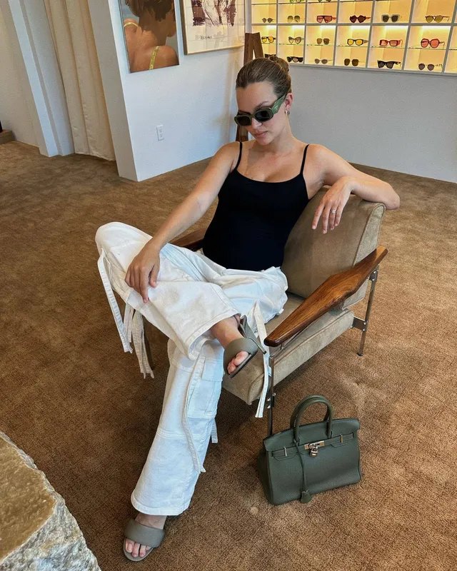 Josephine Skriver sitting in a chair wearing black flat sandals and white trousers.