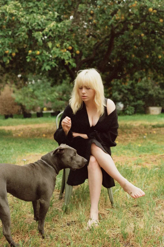 Billie Eilish a black dress while seated beside a dog.