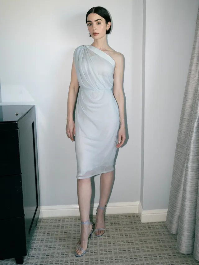 Lily Collins standing barefoot indoors in a pale one-shoulder dress.