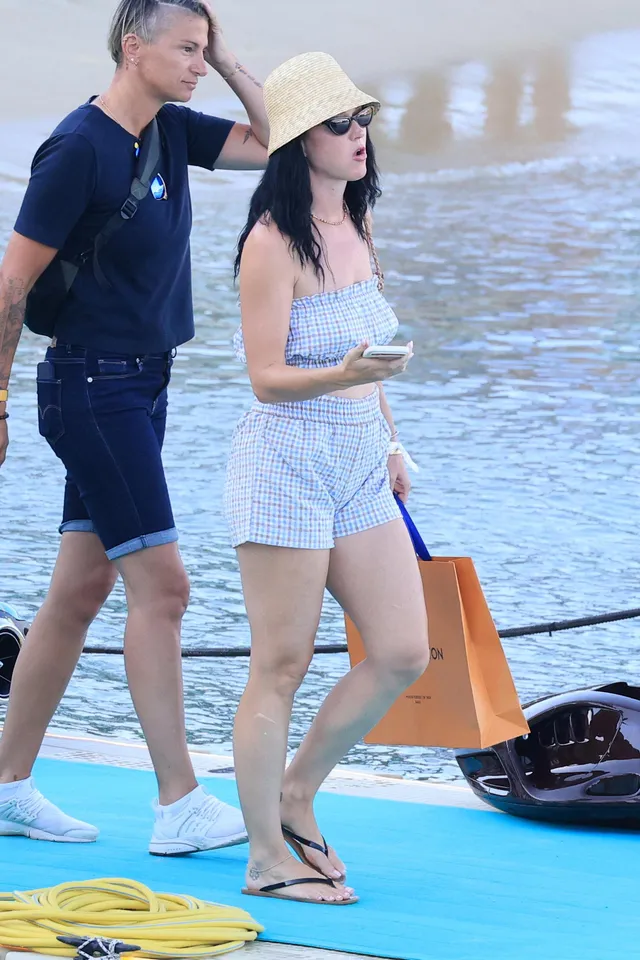 Katy Perry standing beside a boat wearing shorts and flip flops.
