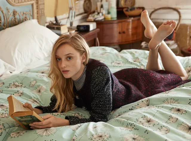 Phoebe Dynevor reading while lying on a patterned bed.