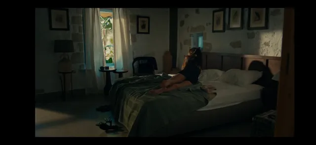 Hande Erçel reclining barefoot on a bed in a dimly lit scene.
