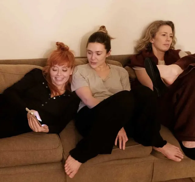 Elizabeth Olsen sitting barefoot between two women on a sofa.
