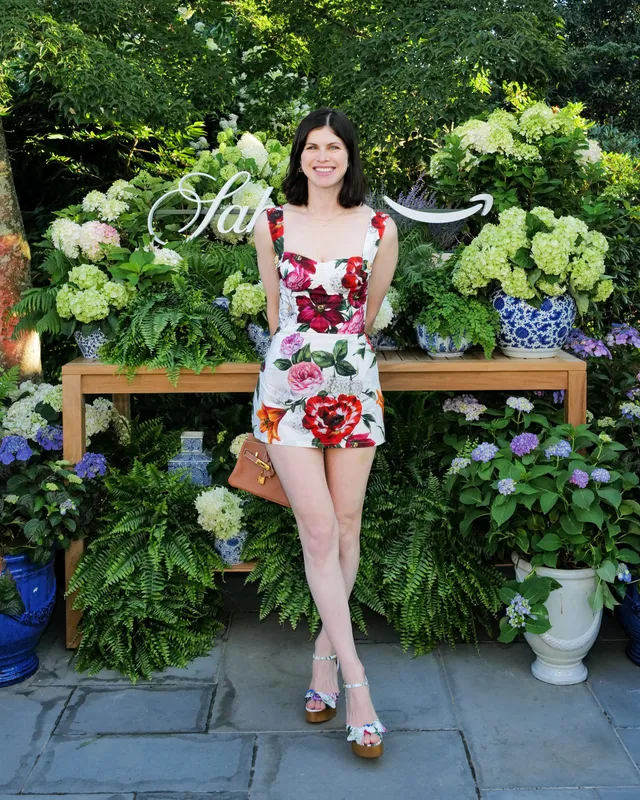 Alexandra Daddario posing beside flowers in a floral dress and strappy heels.