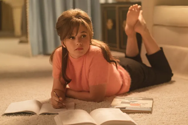 Cailee Spaeny lying barefoot on a carpet while reading in a film scene.