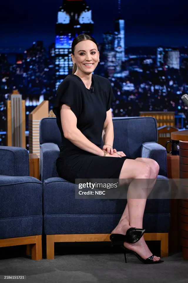 Kaley Cuoco sitting on a television set in a black dress and heels.