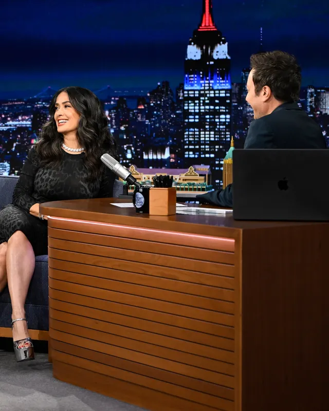Salma Hayek seated beside a talk show desk in a black dress.